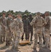 A group of military personnel in camouflage uniforms are gathered in an outdoor arid training environment, appearing to conduct a briefing or instruction session. Two individuals in the center wear distinctive darker camouflage uniforms with body armor and helmets, suggesting they may be from a different military force than the others. Artillery shells or large munitions are visible on the ground in the foreground.