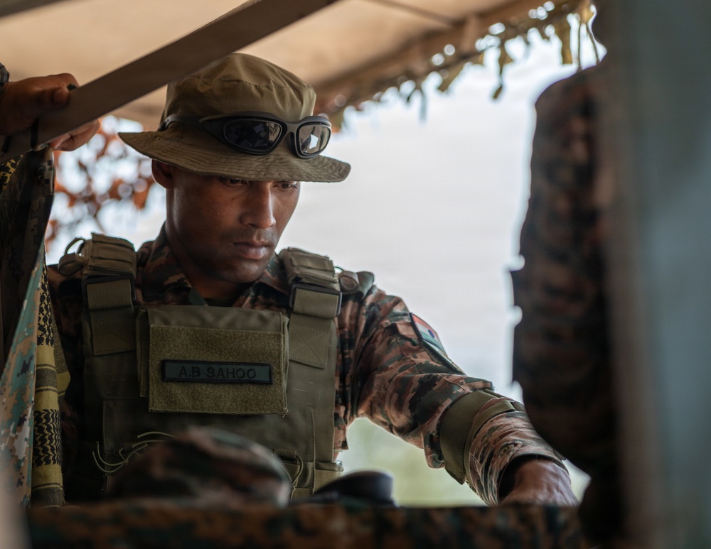 A military soldier identified by a name tape reading 'A.B. SAHOO' is wearing tactical gear including a plate carrier vest, camouflage uniform, a boonie hat, and protective goggles resting on the brim. The soldier appears to be operating within or near a covered position, possibly a field observation post or tactical shelter with camouflage netting visible. The image is a close-up, professionally composed photograph with strong depth of field.