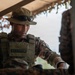 A military soldier identified by a name tape reading 'A.B. SAHOO' is wearing tactical gear including a plate carrier vest, camouflage uniform, a boonie hat, and protective goggles resting on the brim. The soldier appears to be operating within or near a covered position, possibly a field observation post or tactical shelter with camouflage netting visible. The image is a close-up, professionally composed photograph with strong depth of field.