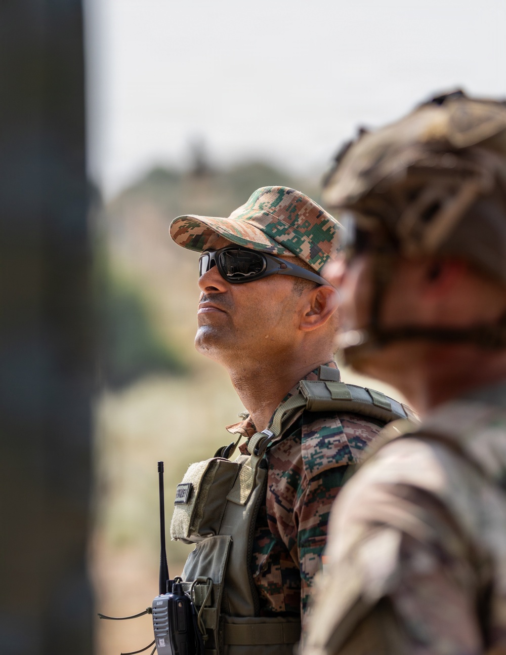 The image shows two military personnel in the foreground, with one sharply in focus wearing digital camouflage uniform and tactical gear. The in-focus soldier is wearing a camouflage patrol cap, tactical sunglasses, a plate carrier vest, and carries a handheld radio. A second soldier is visible in the blurred foreground wearing a camouflage helmet.