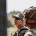 The image shows two military personnel in the foreground, with one sharply in focus wearing digital camouflage uniform and tactical gear. The in-focus soldier is wearing a camouflage patrol cap, tactical sunglasses, a plate carrier vest, and carries a handheld radio. A second soldier is visible in the blurred foreground wearing a camouflage helmet.