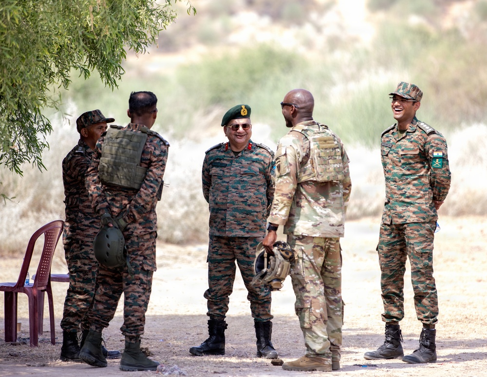 A group of five military personnel in various uniforms are gathered outdoors in an arid, semi-vegetated area, appearing to engage in a relaxed conversation. The individuals wear distinctly different camouflage patterns and insignia, suggesting a multinational military meeting or joint exercise. The atmosphere appears informal and collegial, with visible smiling among the participants.