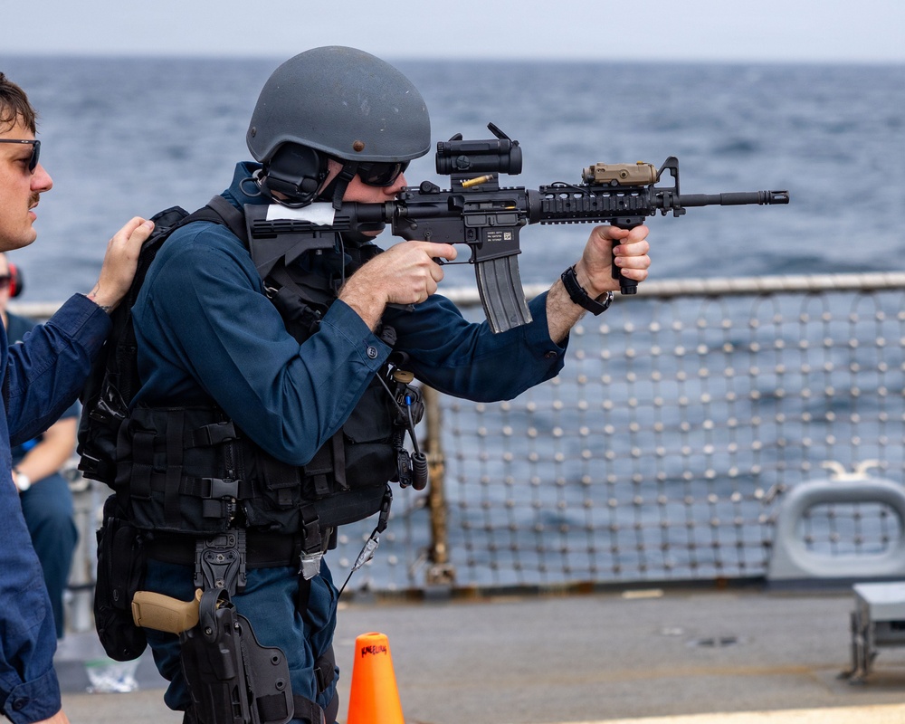 A military or law enforcement personnel in tactical gear is aiming an M4-style carbine rifle on what appears to be the deck of a naval vessel at sea. The individual is wearing a ballistic helmet, body armor/plate carrier, and has a sidearm holstered on the hip. A second person in civilian-style clothing is visible to the left, appearing to observe or assist.