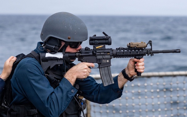 A military or law enforcement personnel in tactical gear is aiming an M4-style carbine rifle on what appears to be the deck of a naval vessel at sea. The individual is wearing a ballistic helmet, body armor/plate carrier, and has a sidearm holstered on the hip. A second person in civilian-style clothing is visible to the left, appearing to observe or assist.