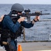 A military or law enforcement personnel in tactical gear is aiming an M4-style carbine rifle on what appears to be the deck of a naval vessel at sea. The individual is wearing a ballistic helmet, body armor/plate carrier, and has a sidearm holstered on the hip. A second person in civilian-style clothing is visible to the left, appearing to observe or assist.