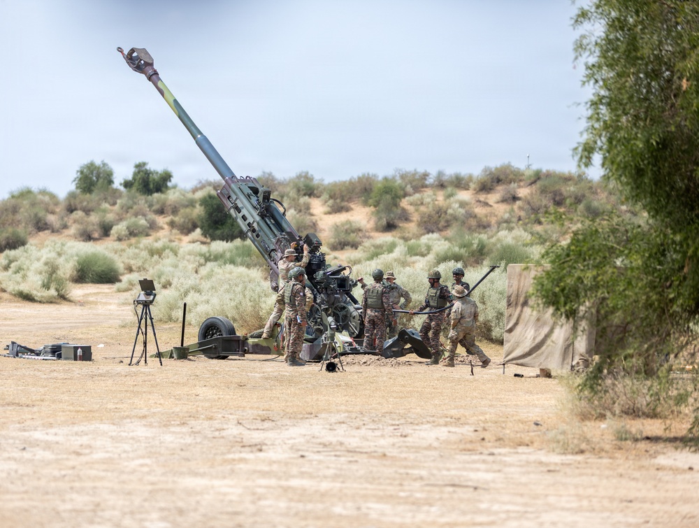 A group of military personnel in camouflage uniforms are operating a large artillery piece, likely an M777 howitzer or similar towed howitzer, in an arid desert training environment. The barrel is elevated at a high angle, suggesting the crew is in the process of loading or preparing to fire. The setting appears to be a military training range based on the open terrain and supporting equipment visible nearby.