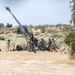 A group of military personnel in camouflage uniforms are operating a large artillery piece, likely an M777 howitzer or similar towed howitzer, in an arid desert training environment. The barrel is elevated at a high angle, suggesting the crew is in the process of loading or preparing to fire. The setting appears to be a military training range based on the open terrain and supporting equipment visible nearby.