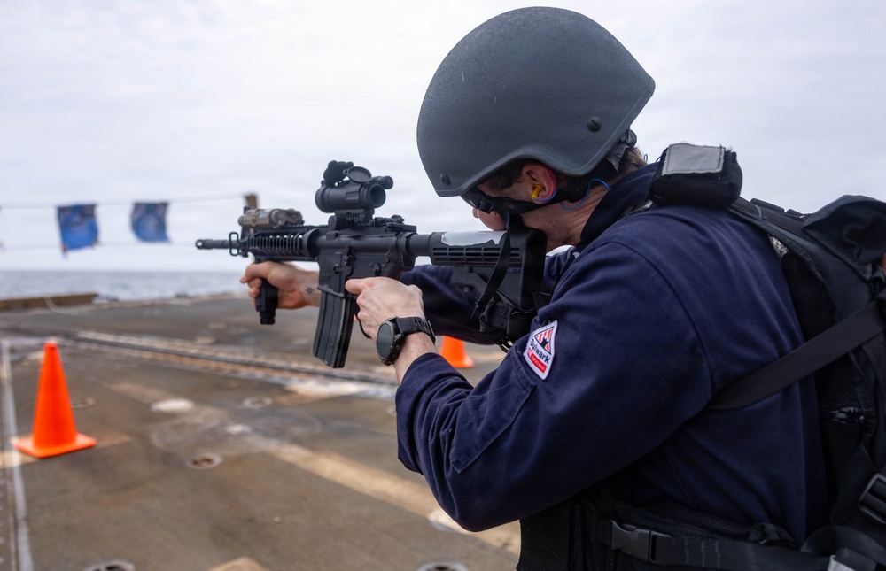 A military or law enforcement personnel is aiming an assault rifle on what appears to be the deck of a naval vessel. The individual is wearing full tactical gear including a ballistic helmet, ear protection, and a dark blue uniform with a visible patch. Orange traffic cones and blue pennant flags are visible in the background, suggesting a training exercise or qualification drill.