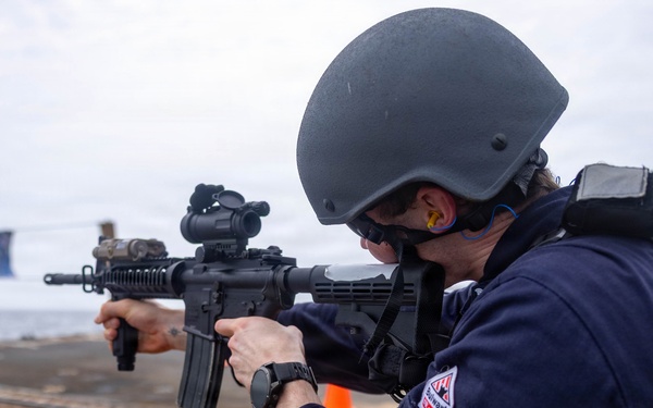 A military or law enforcement personnel is aiming an assault rifle on what appears to be the deck of a naval vessel. The individual is wearing full tactical gear including a ballistic helmet, ear protection, and a dark blue uniform with a visible patch. Orange traffic cones and blue pennant flags are visible in the background, suggesting a training exercise or qualification drill.