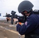 A military or law enforcement personnel is aiming an assault rifle on what appears to be the deck of a naval vessel. The individual is wearing full tactical gear including a ballistic helmet, ear protection, and a dark blue uniform with a visible patch. Orange traffic cones and blue pennant flags are visible in the background, suggesting a training exercise or qualification drill.