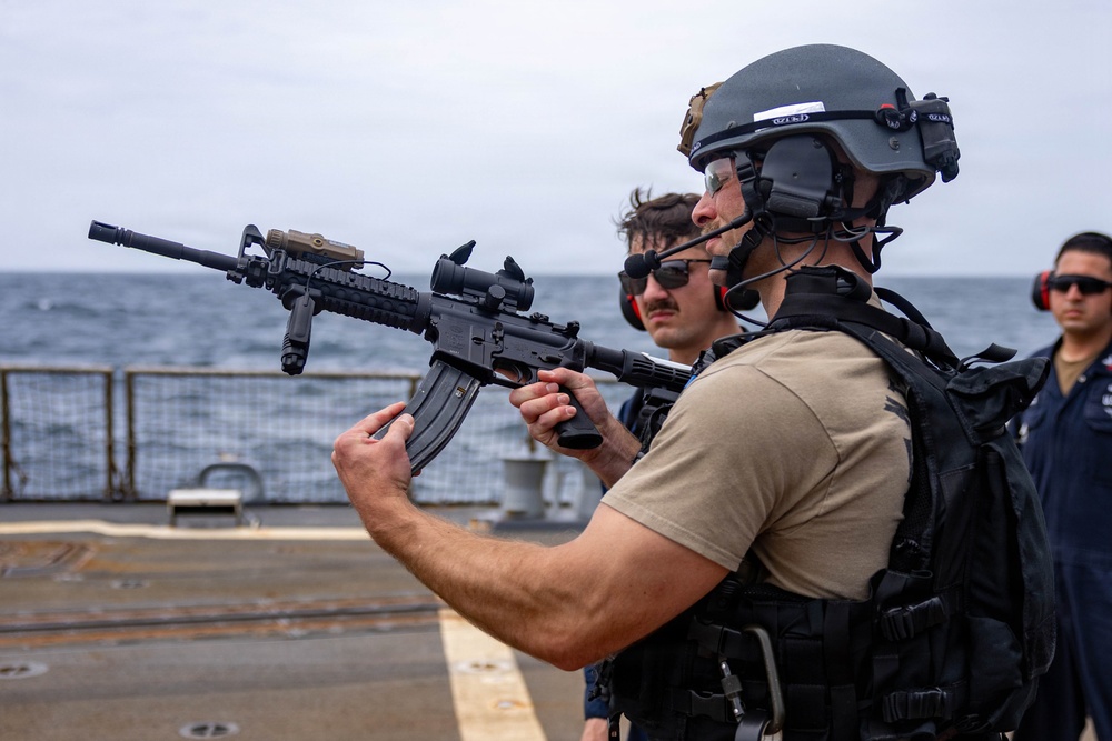 A tactical operator in full combat gear holds an M4-style carbine rifle on what appears to be the deck of a naval vessel at sea. The individual is equipped with a ballistic helmet, communications headset, body armor, and tactical vest. Additional personnel in the background appear to be observing or participating in a training or security exercise.