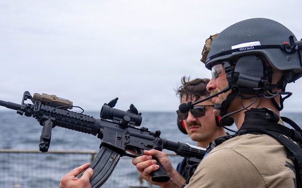 A tactical operator in full combat gear holds an M4-style carbine rifle on what appears to be the deck of a naval vessel at sea. The individual is equipped with a ballistic helmet, communications headset, body armor, and tactical vest. Additional personnel in the background appear to be observing or participating in a training or security exercise.