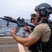 A tactical operator in full combat gear holds an M4-style carbine rifle on what appears to be the deck of a naval vessel at sea. The individual is equipped with a ballistic helmet, communications headset, body armor, and tactical vest. Additional personnel in the background appear to be observing or participating in a training or security exercise.