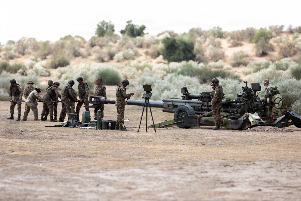A group of military personnel in camouflage uniforms are operating and preparing what appears to be a large artillery piece in an arid, semi-desert training environment. Multiple soldiers are working together to handle a long artillery barrel or projectile, while others attend to equipment around the wheeled howitzer. The setting appears to be a military training exercise based on the organized, deliberate nature of the activity.