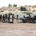 A group of military personnel in camouflage uniforms are operating and preparing what appears to be a large artillery piece in an arid, semi-desert training environment. Multiple soldiers are working together to handle a long artillery barrel or projectile, while others attend to equipment around the wheeled howitzer. The setting appears to be a military training exercise based on the organized, deliberate nature of the activity.