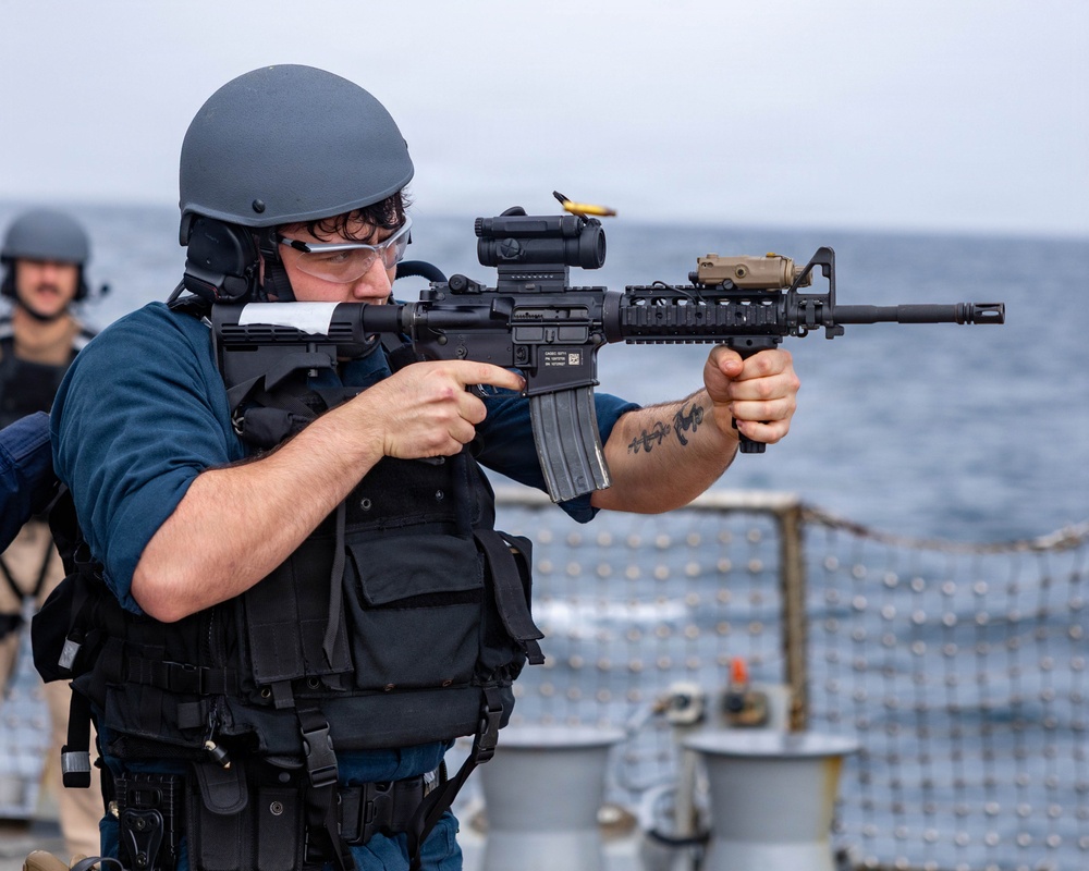 A military or naval personnel member is firing a rifle on the deck of a naval vessel at sea. The shooter is equipped with tactical gear including a ballistic helmet, body armor vest, and eye protection. A second personnel member is visible in the background, also wearing a helmet.