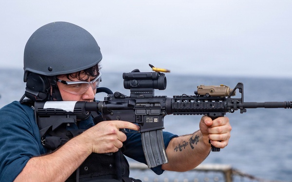 A military or naval personnel member is firing a rifle on the deck of a naval vessel at sea. The shooter is equipped with tactical gear including a ballistic helmet, body armor vest, and eye protection. A second personnel member is visible in the background, also wearing a helmet.