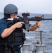 A military or naval personnel member is firing a rifle on the deck of a naval vessel at sea. The shooter is equipped with tactical gear including a ballistic helmet, body armor vest, and eye protection. A second personnel member is visible in the background, also wearing a helmet.