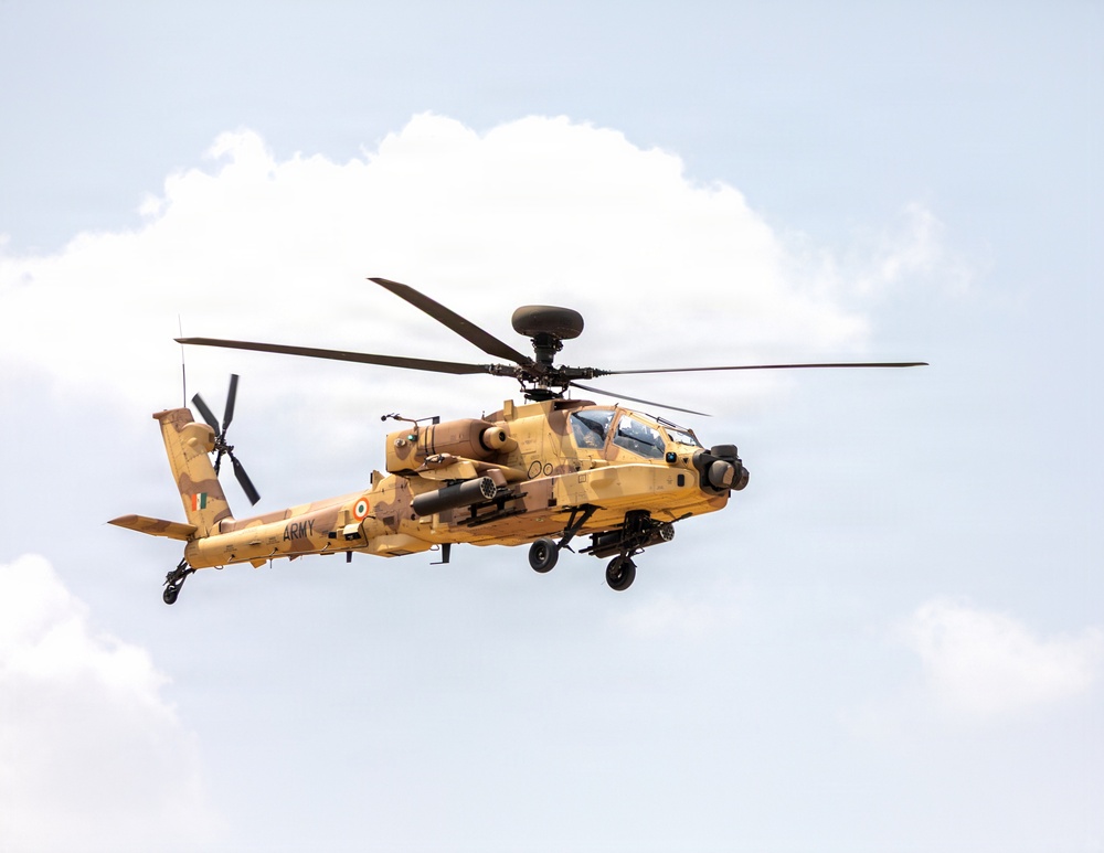 A military attack helicopter painted in desert tan/sand camouflage is captured in flight against a partly cloudy sky. The aircraft bears the word 'ARMY' on its fuselage along with what appears to be an Indian national roundel (tricolor cockade), identifying it as belonging to the Indian Army. The helicopter is equipped with weapons systems and sensor pods consistent with an AH-64 Apache attack helicopter configuration.