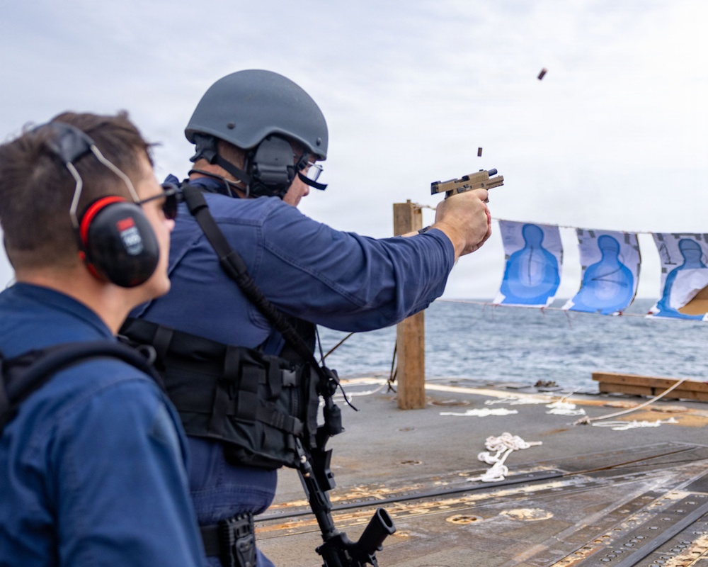 A military or law enforcement personnel is conducting a live-fire pistol qualification exercise on the deck of a naval vessel at sea. A spent brass casing is visibly ejecting from the tan/FDE-colored semi-automatic pistol. A second person wearing red ear protection observes from the left foreground.
