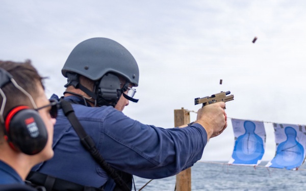 A military or law enforcement personnel is conducting a live-fire pistol qualification exercise on the deck of a naval vessel at sea. A spent brass casing is visibly ejecting from the tan/FDE-colored semi-automatic pistol. A second person wearing red ear protection observes from the left foreground.