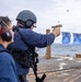 A military or law enforcement personnel is conducting a live-fire pistol qualification exercise on the deck of a naval vessel at sea. A spent brass casing is visibly ejecting from the tan/FDE-colored semi-automatic pistol. A second person wearing red ear protection observes from the left foreground.