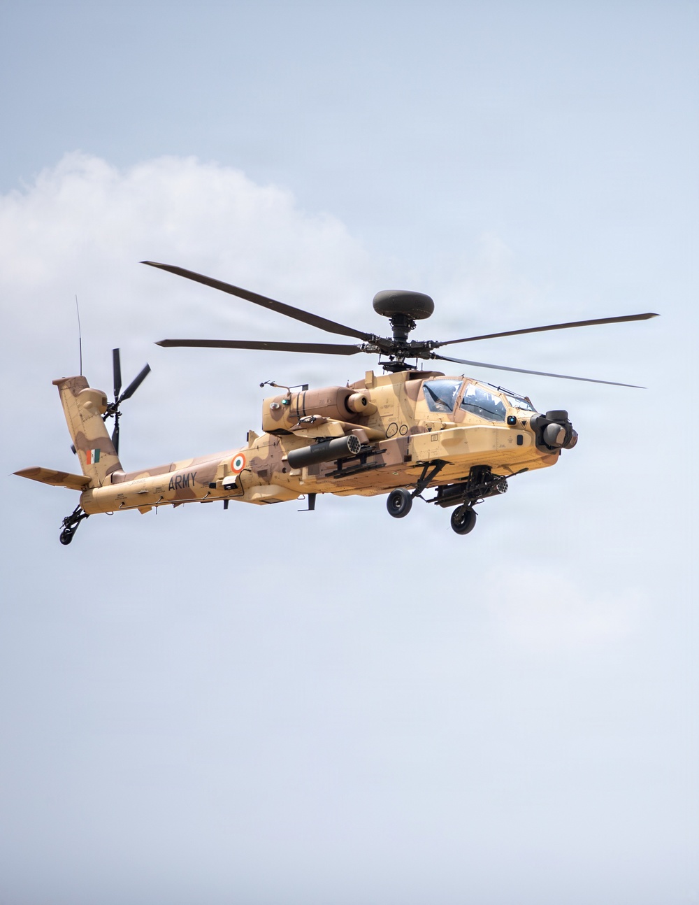 A military attack helicopter is captured in flight against a light blue sky with partial cloud cover. The aircraft displays desert camouflage livery in tan and brown tones, with visible Indian Army markings including the Indian tricolor roundel and the word 'ARMY' on the fuselage. The helicopter appears to be an AH-64 Apache variant operated by the Indian Army.
