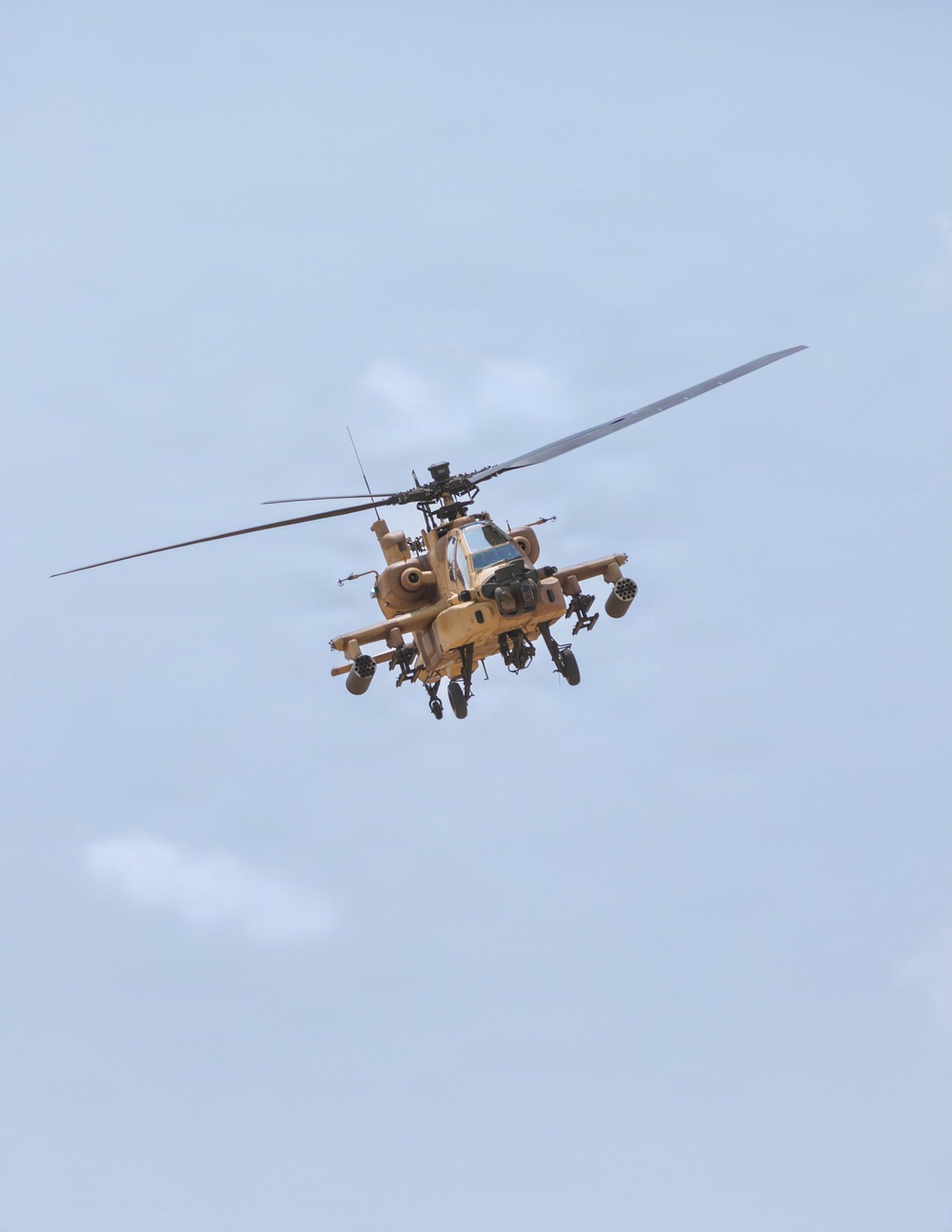A military attack helicopter is photographed in flight from a low angle against an overcast sky. The aircraft is painted in a desert tan/sand camouflage scheme and is fully armed with visible weapons systems. The helicopter appears to be an AH-64 Apache or similar attack variant based on its distinctive tandem cockpit and weapons configuration.