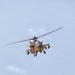 A military attack helicopter is photographed in flight from a low angle against an overcast sky. The aircraft is painted in a desert tan/sand camouflage scheme and is fully armed with visible weapons systems. The helicopter appears to be an AH-64 Apache or similar attack variant based on its distinctive tandem cockpit and weapons configuration.