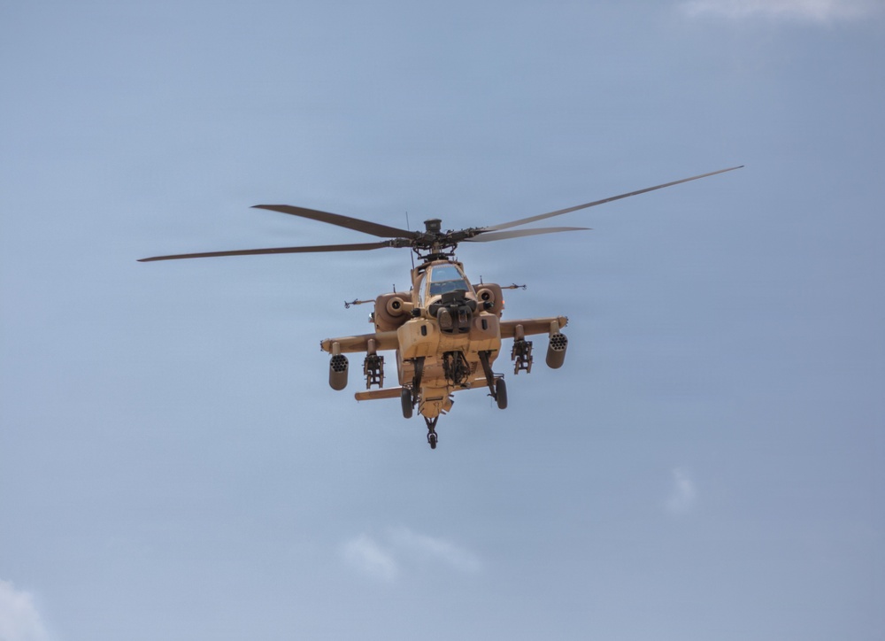 A military attack helicopter is photographed head-on in flight against a partly cloudy blue sky. The aircraft is painted in a desert tan/sand camouflage scheme and is visibly armed with weapons systems on its stub wings. The image captures the helicopter in a forward-facing orientation, providing a clear view of its nose, rotor system, and weapons loadout.