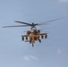 A military attack helicopter is photographed head-on in flight against a partly cloudy blue sky. The aircraft is painted in a desert tan/sand camouflage scheme and is visibly armed with weapons systems on its stub wings. The image captures the helicopter in a forward-facing orientation, providing a clear view of its nose, rotor system, and weapons loadout.
