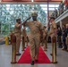 A formal U.S. Navy military ceremony is taking place inside what appears to be an indoor facility, likely a change of command or retirement ceremony. A senior naval officer in khaki service uniform walks along a red carpet flanked by brass stanchions while fellow sailors render salutes. Civilian guests and uniformed personnel line the sides observing the proceedings.