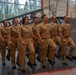 A group of seven U.S. Navy personnel in tan Service Uniform B (SDB) are marching in formation during an official ceremony. The event appears to take place inside a naval facility or museum, with a banner referencing 'USS Whit[ehat]' and the phrase 'Teaching' visible in the background. An American flag and what appears to be a ship's hull or exhibit structure are also visible behind the formation.