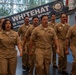 A group of U.S. Navy personnel in tan Service Uniform (Type III) are marching in formation while chanting or reciting in unison. The event appears to be a formal military ceremony, likely a Chief Petty Officer pinning or advancement ceremony, taking place indoors at what appears to be Naval Station Great Lakes. A banner reading 'WHITEHAT' and 'SERVING THOSE WHO SERVE' is visible in the background.
