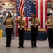 A military color guard ceremony is taking place inside what appears to be a naval facility or armory. Four uniformed personnel in tan/khaki Navy uniforms stand at attention, with two center members carrying flags including the American flag. A senior officer stands at a podium to the left, and a large American flag backdrop decorates the ceremonial stage.