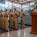 A U.S. Navy military ceremony is taking place inside what appears to be a naval training facility. A senior officer wearing a cover (hat) and service khaki uniform stands at a wooden podium addressing a formation of sailors in service khaki uniforms. A banner referencing 'America's Navy - A Global Force for Good' and a USS vessel name are visible in the background.
