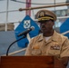 A U.S. Navy officer in service khaki uniform is speaking at a podium with a microphone, appearing to deliver a formal address or speech. The setting appears to be aboard a naval vessel, evidenced by the ship's deck equipment, rigging, and blue ceremonial covers with anchor insignia visible in the background. The officer wears a combination cover with gold insignia, multiple service ribbons, and qualification badges.