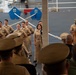 A formal U.S. Navy military ceremony is taking place aboard what appears to be a naval vessel or ship's hangar bay. A senior officer wearing a combination cover with gold braid is speaking at a wooden podium while a group of sailors in khaki service uniforms stand at attention. An audience of uniformed personnel and civilians observes the proceedings, with at least one person recording on a smartphone.
