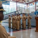 A formal U.S. Navy ceremony is taking place inside a naval facility or museum, with multiple sailors in khaki service uniforms standing in formation. A civilian man in a blue blazer is speaking at a wooden podium, while a senior officer in dress whites observes from the foreground. A banner reading 'WHITEHAT' and an 'America's Navy - A Global Force for Good' sign are visible in the background near what appears to be a naval vessel on display.