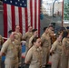 A group of U.S. Navy personnel in tan/khaki service uniforms are standing at attention during a formal military ceremony. A large American flag is prominently displayed in the background, and an officer appears to be speaking at a podium. The setting appears to be inside a naval facility or hangar, with a vessel visible in the background.