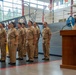A formal U.S. Navy ceremony is taking place inside what appears to be a naval museum or hangar facility, with a vessel visible in the background. A female Navy officer is speaking at a wooden podium while a formation of approximately eight sailors in khaki service uniforms stands at attention to her left. The setting includes nautical displays, flags, and a banner partially visible in the background.