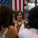 A military promotion or pinning ceremony is taking place in what appears to be a hangar or military facility. A female service member in tan/khaki U.S. Navy uniform is having insignia or rank devices pinned onto her collar by two civilians, likely family members. An American flag is prominently displayed in the background, and another uniformed service member is visible behind the group.