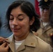 A military promotion or pinning ceremony is taking place, with a person in a tan/khaki uniform having an insignia or rank device pinned onto their collar. The subject appears emotional, with eyes closed and a slight smile, suggesting a meaningful milestone. A second uniformed individual wearing a peaked cap and ribbons is visible in the background.