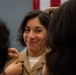 A female military service member in a tan/khaki uniform is smiling as two individuals pin insignia or rank devices onto her collar. An American flag is partially visible in the background, suggesting a formal military ceremony. The subject appears pleased, indicating this may be a promotion or award ceremony.