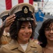 A U.S. Navy service member is having a Chief Petty Officer cover (hat) placed on her head during what appears to be a CPO pinning or advancement ceremony. She is wearing khaki service uniform with visible warfare insignia and collar devices. Two civilians, likely family members, are present and smiling alongside her.