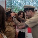 A U.S. Navy chief petty officer pinning ceremony is taking place indoors, likely in a gymnasium or ceremonial hall. A heavily tattooed male sailor in khaki uniform is placing a combination cover (hat) on a female sailor, a traditional part of the CPO pinning/frocking ceremony. Family members, including what appears to be a young woman and a child, are present and participating in the moment.