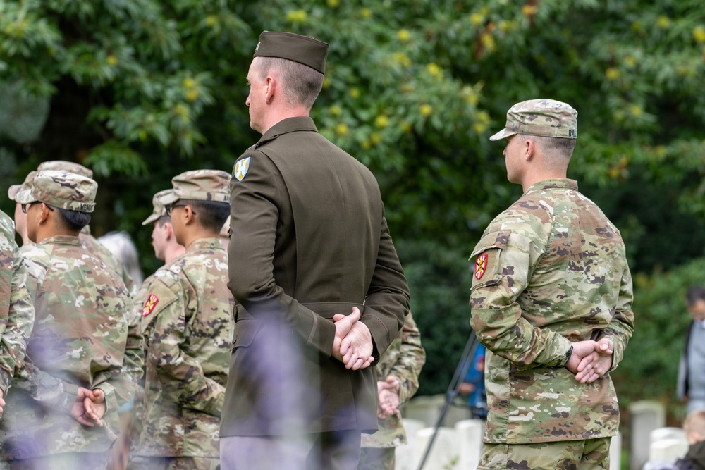 A group of U.S. Army soldiers are standing at attention or in a rest position during what appears to be a formal military ceremony, likely at a cemetery given the visible grave markers in the background. One soldier in the center is wearing the Army Service Uniform (ASU) in olive green, while the others wear Operational Camouflage Pattern (OCP) uniforms. All visible soldiers have their hands clasped behind or in front of them in a formal stance.