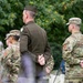 A group of U.S. Army soldiers are standing at attention or in a rest position during what appears to be a formal military ceremony, likely at a cemetery given the visible grave markers in the background. One soldier in the center is wearing the Army Service Uniform (ASU) in olive green, while the others wear Operational Camouflage Pattern (OCP) uniforms. All visible soldiers have their hands clasped behind or in front of them in a formal stance.