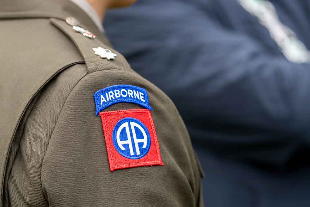 The image shows a close-up of a U.S. Army dress uniform shoulder, featuring the distinctive 82nd Airborne Division patch. The patch consists of a blue 'AIRBORNE' tab above a red square bearing the 'AA' (All American) insignia within a blue circle. Rank insignia, appearing to be a Lieutenant Colonel oak leaf cluster, is also visible on the collar/shoulder area.