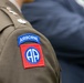 The image shows a close-up of a U.S. Army dress uniform shoulder, featuring the distinctive 82nd Airborne Division patch. The patch consists of a blue 'AIRBORNE' tab above a red square bearing the 'AA' (All American) insignia within a blue circle. Rank insignia, appearing to be a Lieutenant Colonel oak leaf cluster, is also visible on the collar/shoulder area.